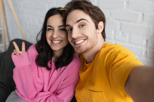 Sorrindo jovem casal sentado no sofá e olhando para a câmera com gesto de vitória na sala de estar — Fotografia de Stock