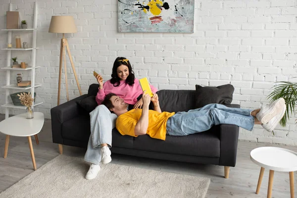 Sonriente joven sentada en el sofá con el teléfono celular cerca de novio acostado con libro en la sala de estar - foto de stock