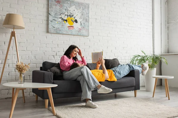 Усміхнена молода жінка сидить на дивані з хлопцем, лежить і читає книгу у вітальні — стокове фото