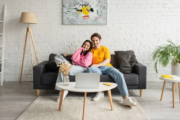 Усміхнена молода пара сидить на дивані і дивиться фільм на ноутбук у вітальні — стокове фото