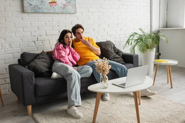 Amazed young couple sitting on couch with open mouths and watching movie on laptop in living room — Stock Photo