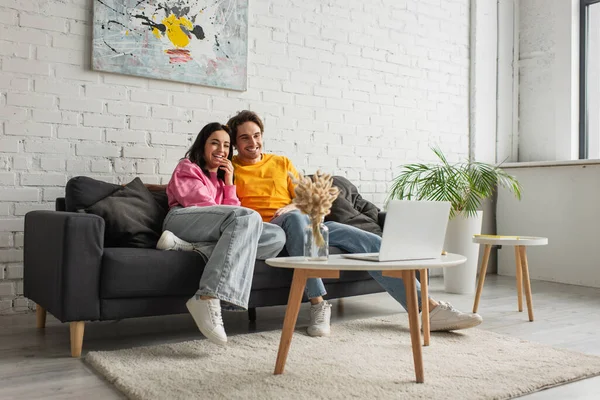 Sonriente joven pareja sentada en el sofá y viendo la película en el ordenador portátil en la sala de estar - foto de stock