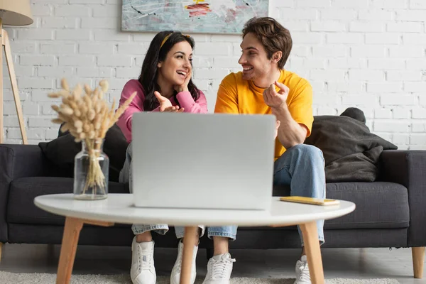 Sonriente joven pareja sentada en el sofá, mirándose entre sí y señalando con los dedos a la computadora portátil en la sala de estar - foto de stock