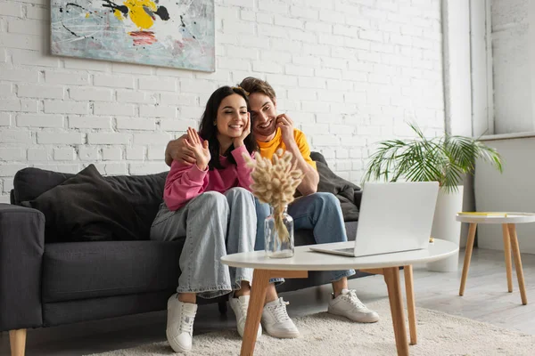 Sonriente mujer joven con la mano agitada sentado con el novio y tener videollamada en el ordenador portátil en la sala de estar - foto de stock