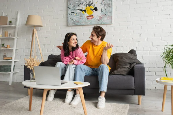 Smiling young couple sitting on couch and gesturing with hands near table with laptop and cellphone in living room — Stock Photo