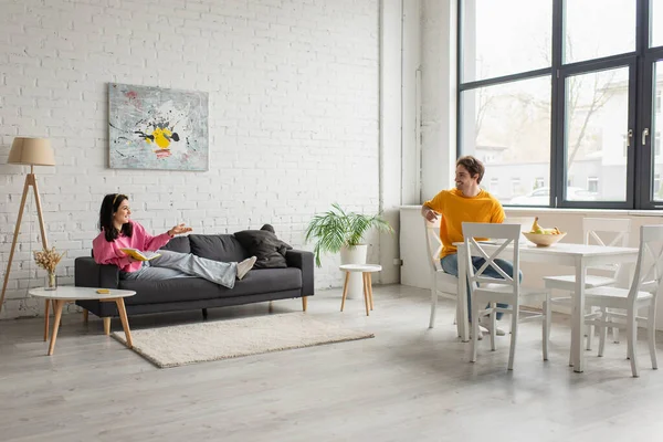 Lächelnde junge Frau liegt mit Buch auf Couch und spricht mit Freund, der mit Laptop im Wohnzimmer am Tisch sitzt — Stockfoto