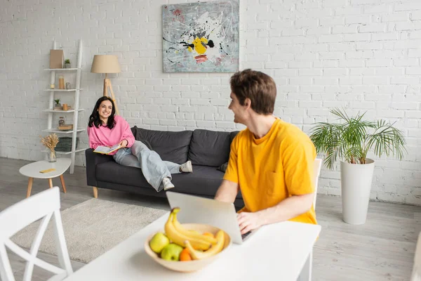 Jeune femme souriante couchée avec livre sur le canapé et regardant petit ami flou assis à la table avec ordinateur portable dans le salon — Stock Photo