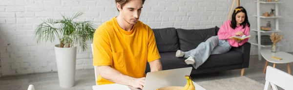 Joven sentado en la mesa con el ordenador portátil cerca de novia borrosa acostado con libro en el sofá en la sala de estar, pancarta — Stock Photo