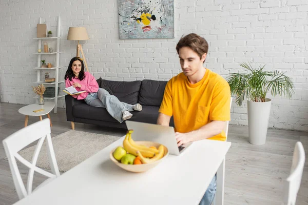 Junge Frau in legerer Kleidung liegt mit Buch auf Couch neben verschwommenem Freund, der mit Laptop im Wohnzimmer am Tisch sitzt — Stockfoto