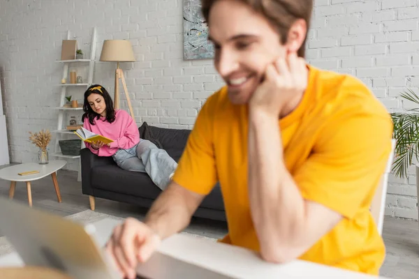 Mujer joven en ropa casual acostado con libro en el sofá cerca borrosa novio sonriente sentado en la mesa con el ordenador portátil en la sala de estar - foto de stock