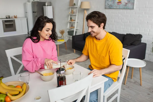 Jeune femme souriante assise à table et versant du lait dans un bol avec des flocons de maïs près du petit ami prenant le petit déjeuner dans le salon — Photo de stock