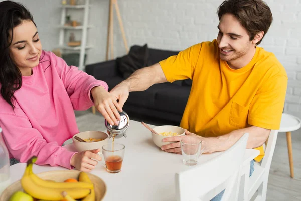 Sonriente joven pareja sentada en la mesa con el desayuno y verter el té de la prensa francesa en la taza en la sala de estar - foto de stock