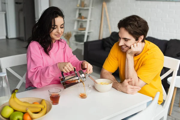Smiling young woman sitting at table near boyfriend with breakfast and pouring tea from french press into cup in living room — Stock Photo