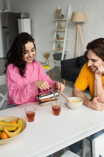 Sorrindo jovem mulher sentada à mesa perto do namorado com café da manhã e derramando chá da imprensa francesa em xícara na sala de estar — Fotografia de Stock