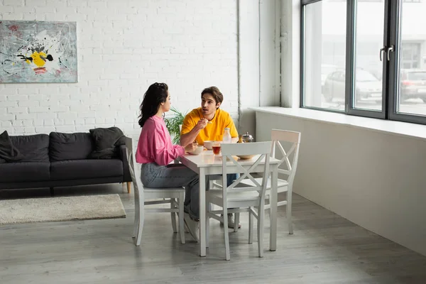 Lächelndes junges Paar sitzt am Tisch und frühstückt im modernen Wohnzimmer — Stockfoto