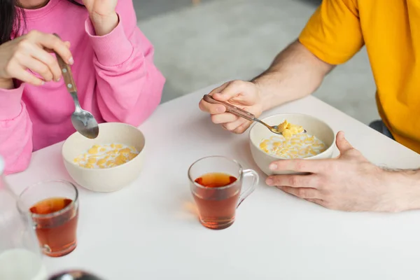 Vista parcial de la pareja joven sentada en la mesa y comiendo hojuelas de maíz cerca de tazas de té en casa - foto de stock