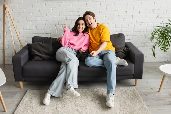 Lächelnder junger Mann zeigt mit Fernbedienung auf Kamera und umarmt Freundin im Wohnzimmer — Stockfoto
