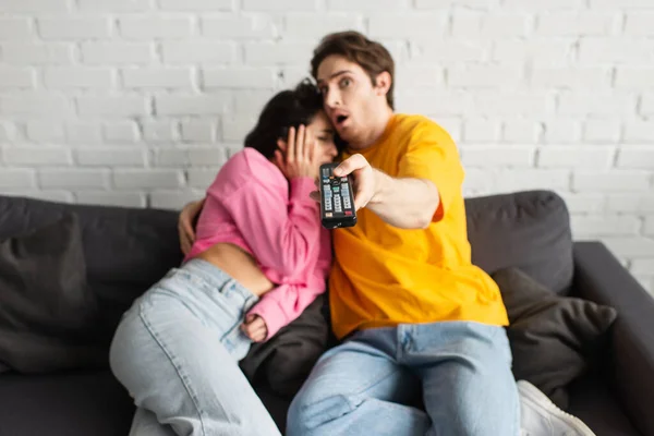 Jovem assustado apontando com controle remoto na câmera e abraçando namorada em casa — Fotografia de Stock