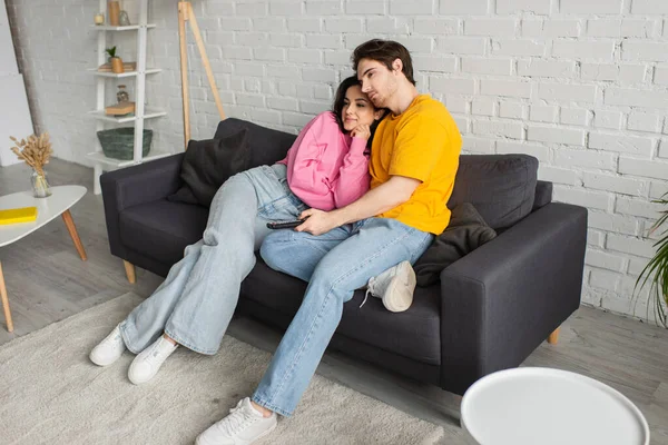 Позитивный молодой человек сидит на диване, держа пульт дистанционного управления и обнимая подругу в гостиной — стоковое фото