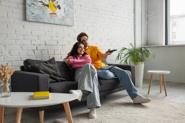 Jovem positivo sentado no sofá, segurando controlador remoto e abraçando namorada na sala de estar moderna — Fotografia de Stock