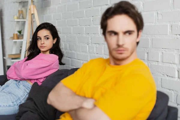 Young woman sitting on couch near blurred disappointed man with crossed arms at home — Stock Photo
