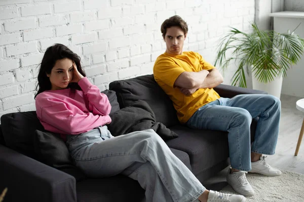 Angry young man with crossed arms sitting on couch and looking at girlfriend with hand near head in living room — Stock Photo