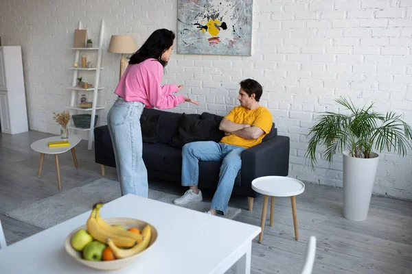 Mujer joven enojada gritando con las manos extendidas cerca de novio sentado en el sofá con los brazos cruzados en la sala de estar - foto de stock