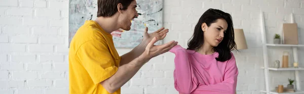 Angry young man yelling with outstretched hands near girlfriend in living room, banner — Stock Photo