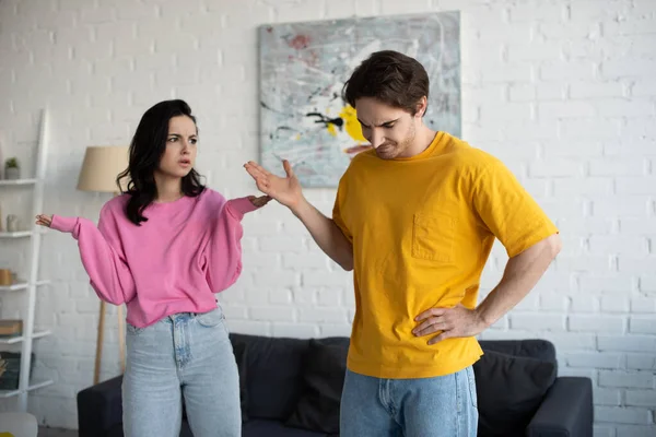 Angry young woman standing with hands in air near boyfriend with refuse gesture in living room — Stock Photo