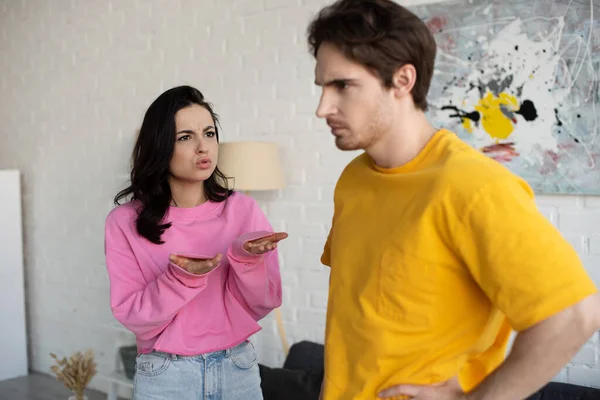 Angry young woman shouting with outstretched hands near boyfriend with hand on hip pose in living room — Stock Photo