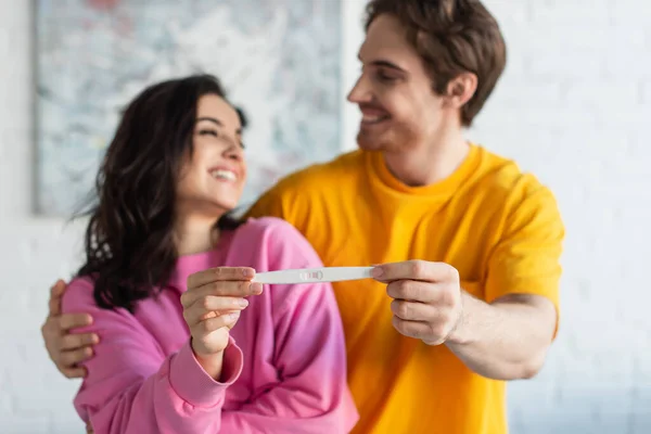 Test di gravidanza in mani di giovane coppia sorridente offuscata che si guarda a casa — Foto stock