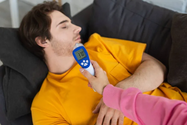 Female hand with non-contact pyrometer near diseased young man lying on couch with closed eyes at home — Stock Photo