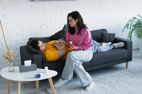 Молодая женщина сидит со стаканом воды и таблетки банка рядом с больным молодым человеком, лежащим на диване с закрытыми глазами дома — стоковое фото
