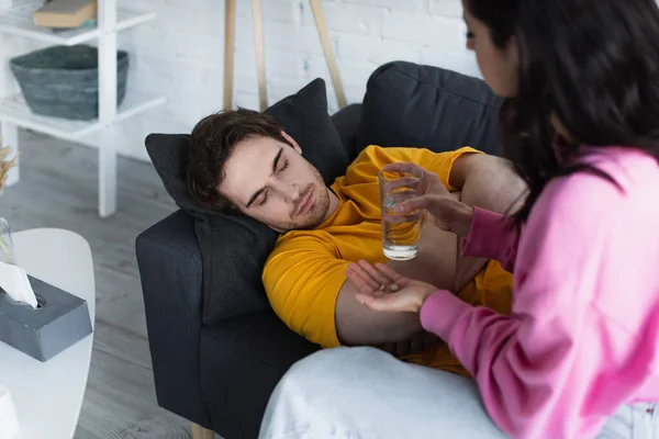 Junge Frau hält Glas Wasser und Tabletten neben kranken jungen Mann, der zu Hause mit geschlossenen Augen auf Couch liegt — Stockfoto