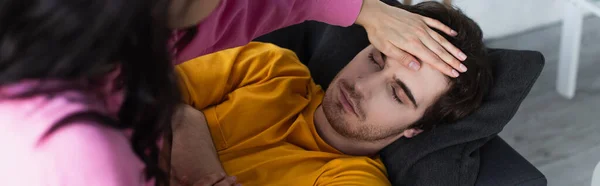 Junge Frau misst Temperatur mit Handfläche auf der Stirn eines kranken jungen Mannes, der zu Hause mit geschlossenen Augen auf dem Sofa liegt, Banner — Stockfoto