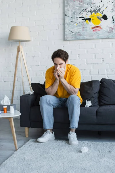 Malato giovane seduto sul divano e soffiando naso con tovagliolo di carta a casa — Foto stock