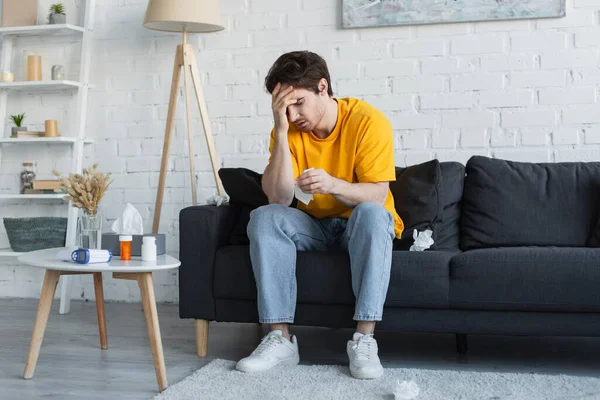 Kranker junger Mann sitzt mit Hand am Kopf auf Couch und hält Papierserviette zu Hause — Stockfoto