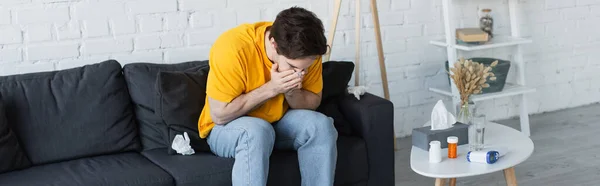 Joven enfermo sentado en el sofá y estornudando con las manos cubriendo la cara en casa, pancarta - foto de stock