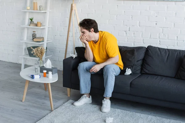 Kranker junger Mann sitzt auf Couch und hustet zu Hause mit der Hand in der Nähe des Gesichts — Stockfoto