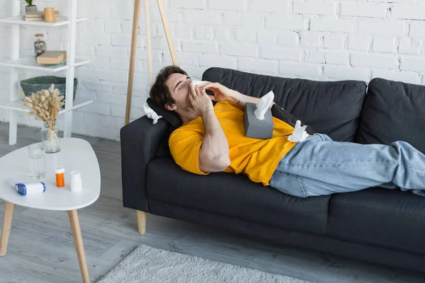 Sick young man lying on couch and blowing nose with paper napkin at home — Stock Photo