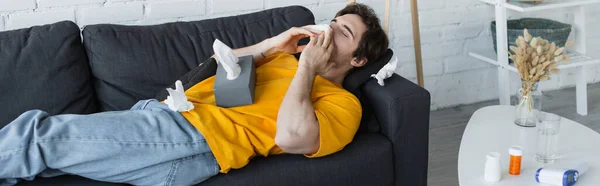 Больной молодой человек лежит на диване и высморкается бумажной салфеткой дома, баннер — стоковое фото