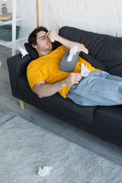 Jeune homme malade couché sur le canapé avec la main près de la tête et tenant une serviette en papier à la maison — Photo de stock