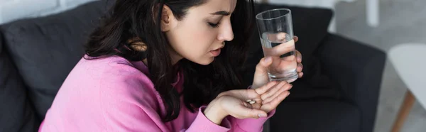 Хвора молода жінка сидить на дивані зі склянкою води і таблеток вдома, банер — стокове фото