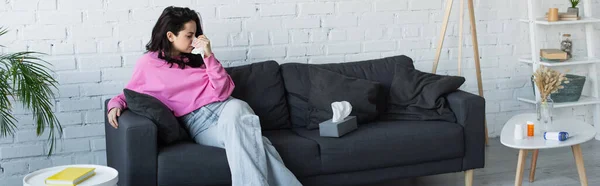 Diseased young woman sitting on couch and blowing nose with paper napkin in living room, banner — Stock Photo