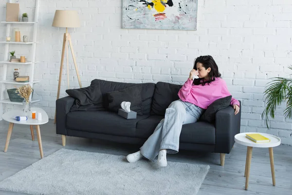 Diseased young woman sitting on couch and blowing nose with paper napkin in living room — Stock Photo