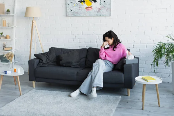 Diseased young woman sitting on couch and blowing nose with paper napkin at home — Stock Photo