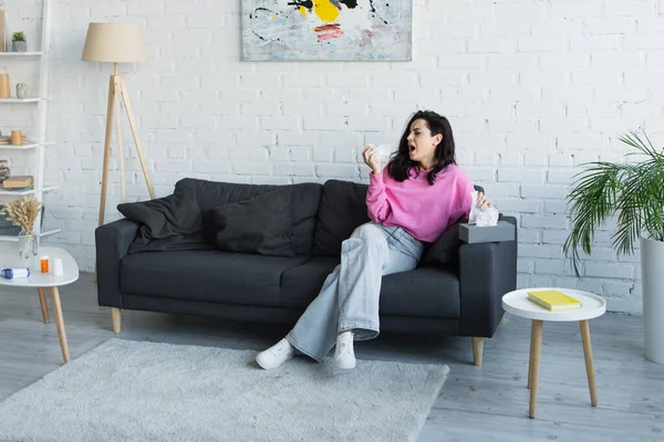 Diseased young woman sitting on couch and coughing with paper napkin in hand at home — Stock Photo