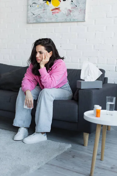 Sick young woman sitting on couch with hand near face and holding paper napkin in living room — Stock Photo