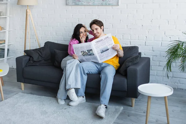 Sorrindo jovem casal sentado no sofá e segurando jornal em casa — Fotografia de Stock