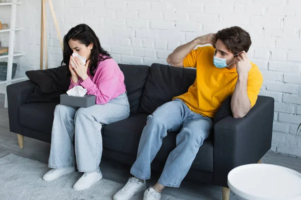 Mujer joven enferma sentada en el sofá y sonándose la nariz con servilleta de papel cerca de novio en máscara médica en la sala de estar - foto de stock